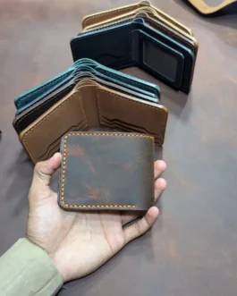 A person holding a closed distressed dark brown handmade leather bifold wallet with yellow stitching, with stacked leather wallets in the background.
